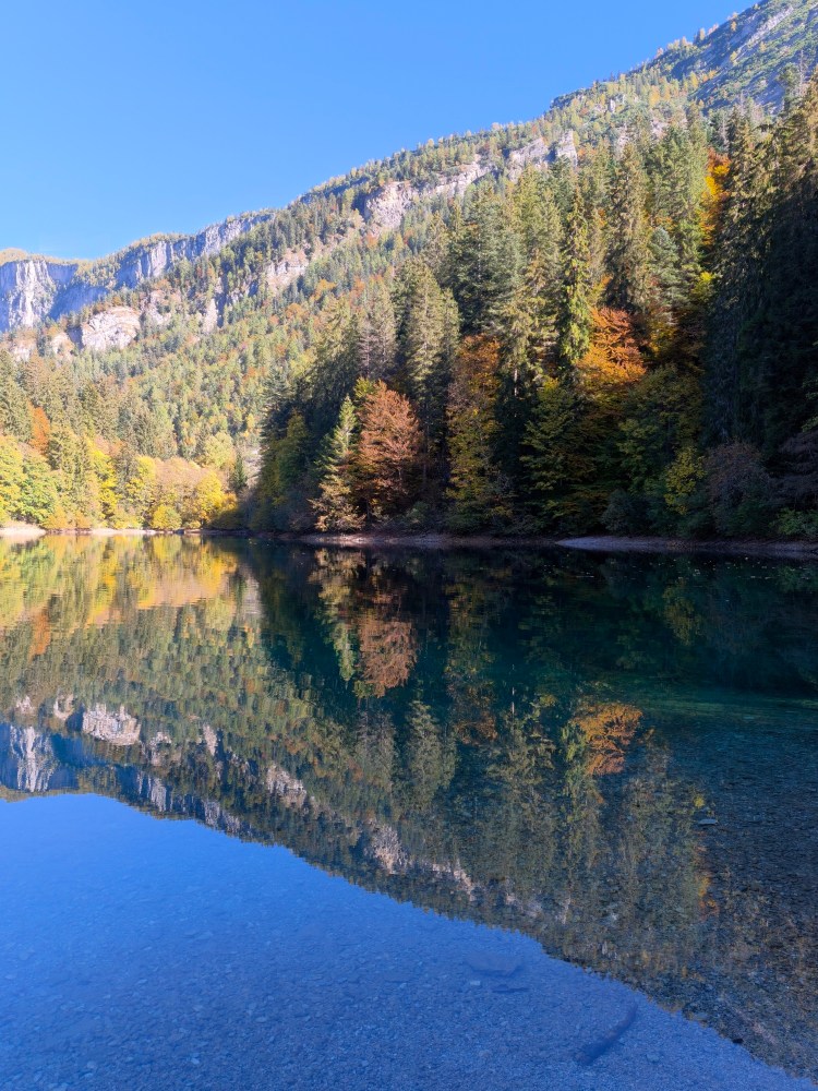 Panorama di un lago circondato da alberi e montagne, con riflessi colorati sulla superficie dell'acqua in una giornata di sole.