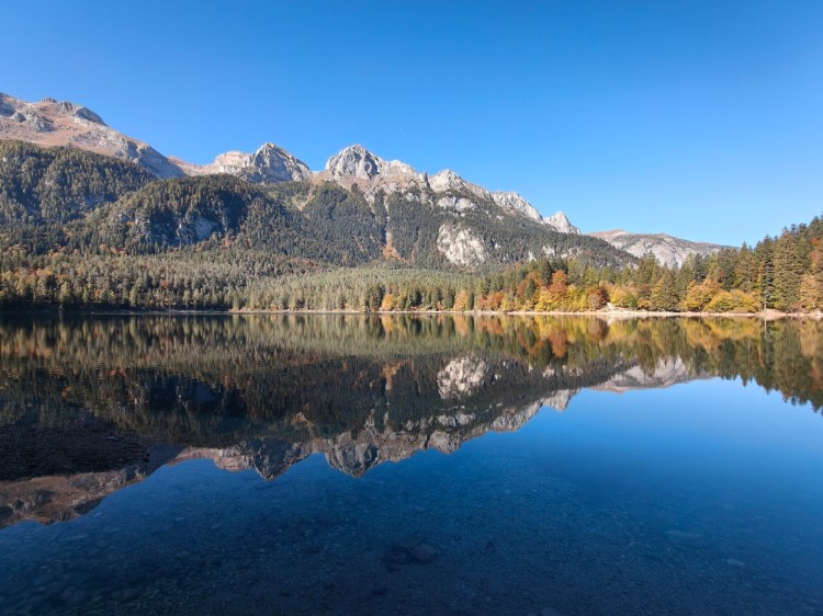 Lago tranquillo circondato da montagne, con riflessi nelle acque cristalline e un cielo azzurro.