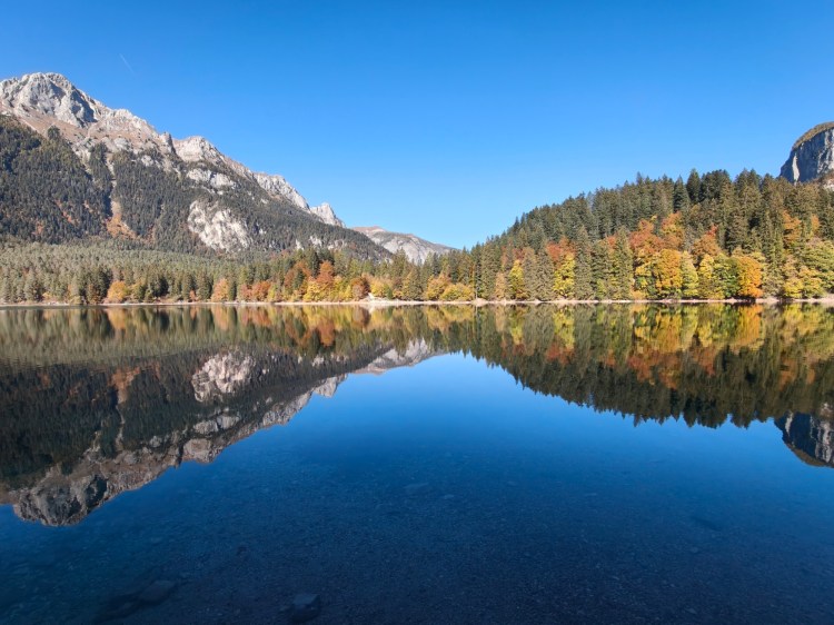 Un lago tranquillo circondato da alberi autunnali e montagne, riflesso nell'acqua, sotto un cielo blu.