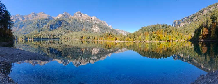Panorama di un lago circondato da montagne e foreste verdi, con un cielo blu limpido e riflessi sull'acqua calma.