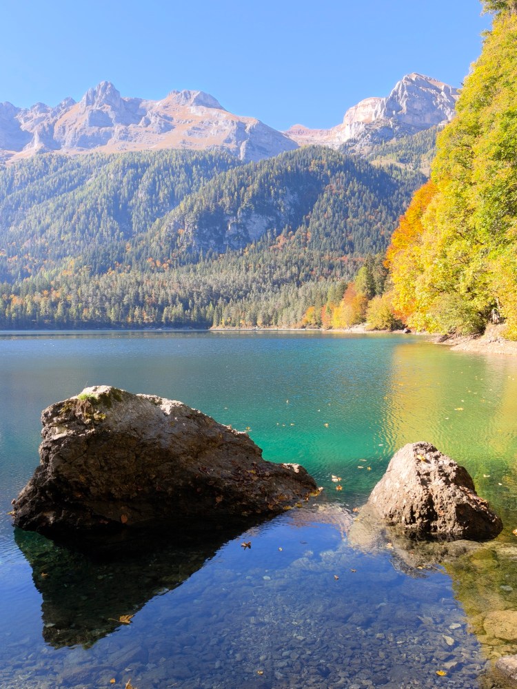 Vista di un lago circondato da montagne e alberi autunnali, con due rocce visibili in primo piano.