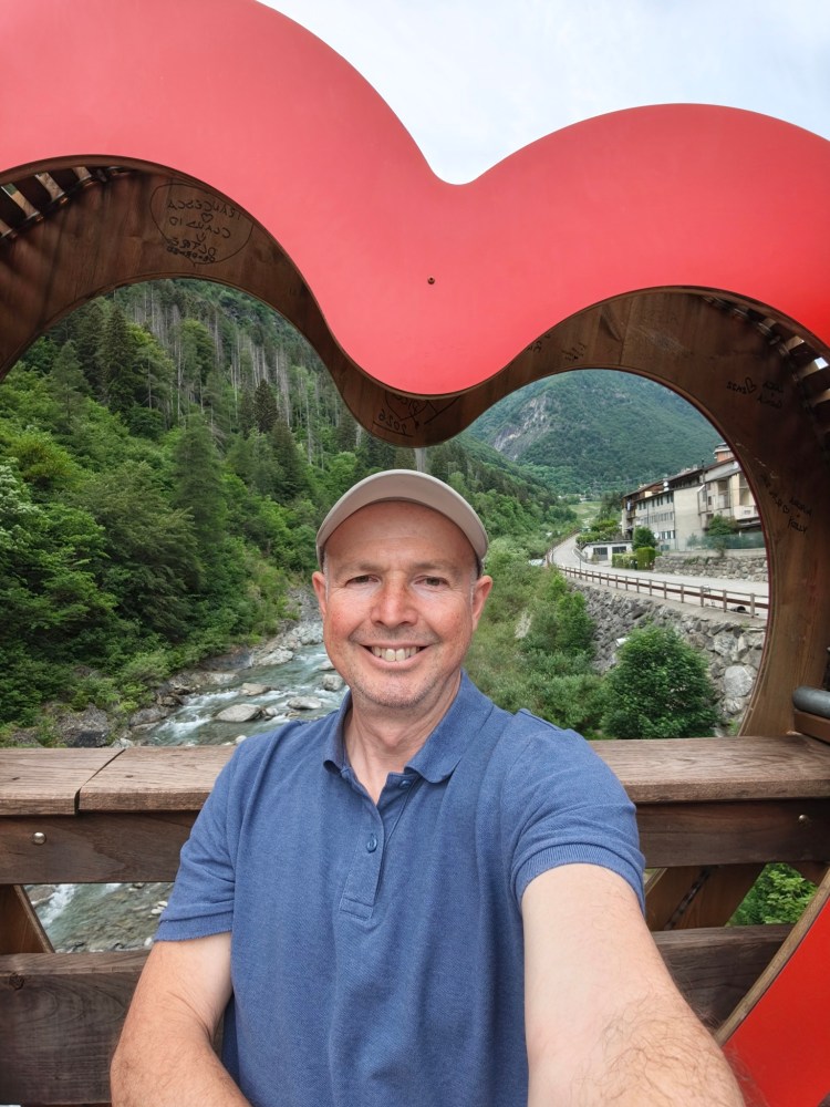 Un uomo sorridente si scatta un selfie attraverso una cornice a forma di cuore, circondato da un paesaggio verde e montano.
